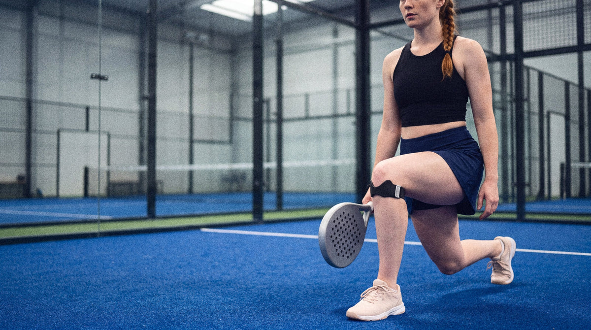 A padel player doing a lunge warm-up beside the glass, strap below her knee