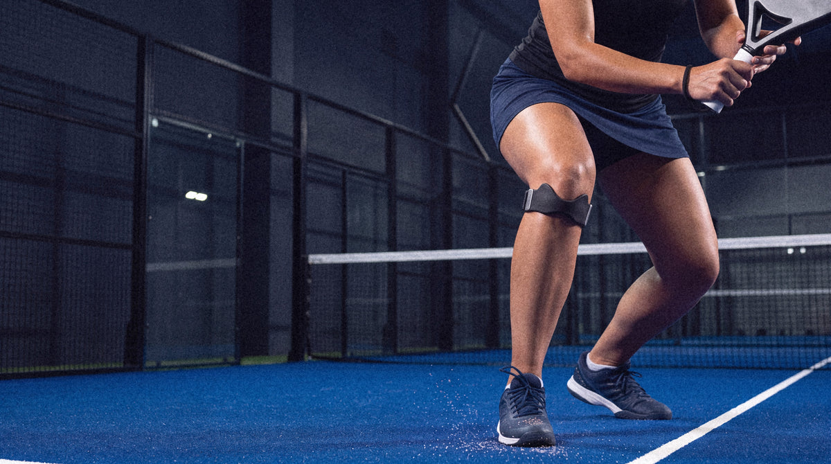 A padel player in a low ready stance behind the net at night, the Second Set strap just below her kneecap
